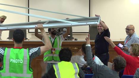 Bevor die Pressekonferenz nach dem Sieg über den Oman losgehen kann, greift Hansi Flick selbst mit an und hilft beim Aufbau der Sponsorenwand. Dazu macht er mit einheimischen Journalisten Selfies.