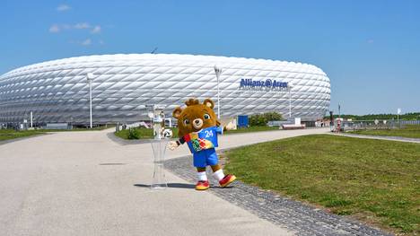 In der Allianz-Arena finden neben dem Eröffnungsspiel weitere fünf Spiele der EM 2024 statt. Maskottchen Albärt präsentiert den Pokal vor der München Fußball Arena
