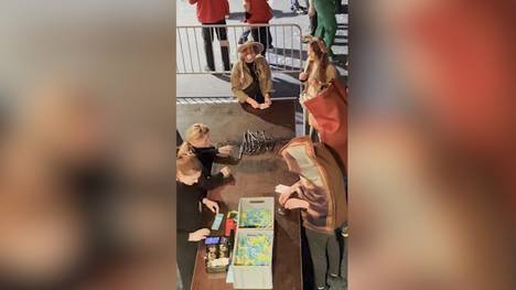 Zwei Darts-Fans möchten beim German Darts Grand Prix in München ihre Jacken abgeben und kommen an ihre Grenzen. Ihre kuriosen Kostüme machen ihnen fast einen Strich durch die Rechnung.