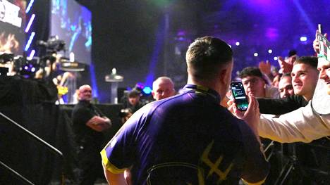 Bei der Premier League of Darts genießt Luke Littler seinen Walk-On und öffnet währenddessen auf dem Handy eines Fans ein FIFA-Pack.