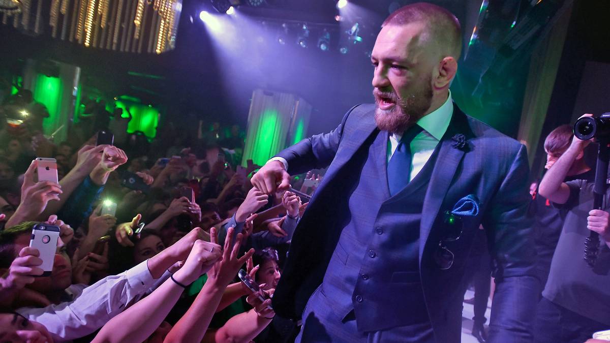 ...und grüßt, während er mit der Linken ein Glas hält, mit den Fingerknöcheln seiner rechten Hand