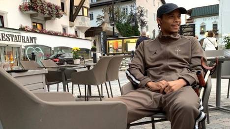 Leroy Sané sitzt im braunen Trainingsanzug lächelnd vor einem Restaurant